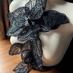Pode incluir: Aplique floral preto com detalhes intrincados de contas e lantejoulas. O aplique apresenta uma flor grande com múltiplas pétalas e folhas delicadas, tudo em uma cor escura. Cristais transparentes estão no centro da flor.