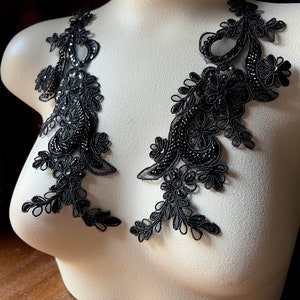 May include: Two black lace appliques with beaded embellishments. The appliques are shaped like a floral design and are displayed on a white mannequin.
