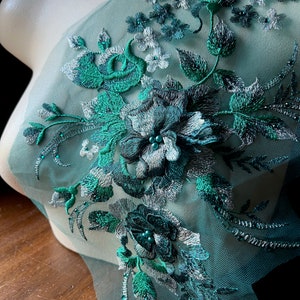 Op de afbeelding: Tealgroen mesh-stof met geborduurde bloemenapplicatie. De applicatie heeft zilveren en groene bloemen met groene bladeren en glinsterende kralen.