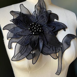 Puede incluir: Una gran aplicación floral negra con pétalos en capas y un centro con cuentas. La aplicación está hecha de tela transparente y está adherida a un maniquí. El diseño presenta costuras intrincadas y una estética delicada y elegante.