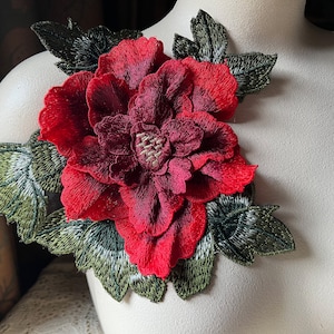 Pode incluir: Uma grande flor vermelha bordada com folhas verdes. A flor é feita de tecido e tem um aspecto texturizado em 3D.
