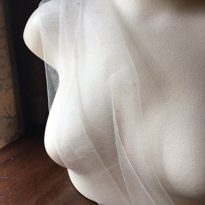 May include: A white tulle fabric draped over a mannequin bust. The tulle is sheer and delicate, with a fine mesh weave. The mannequin is a neutral off-white color, and the background is a dark wood surface.