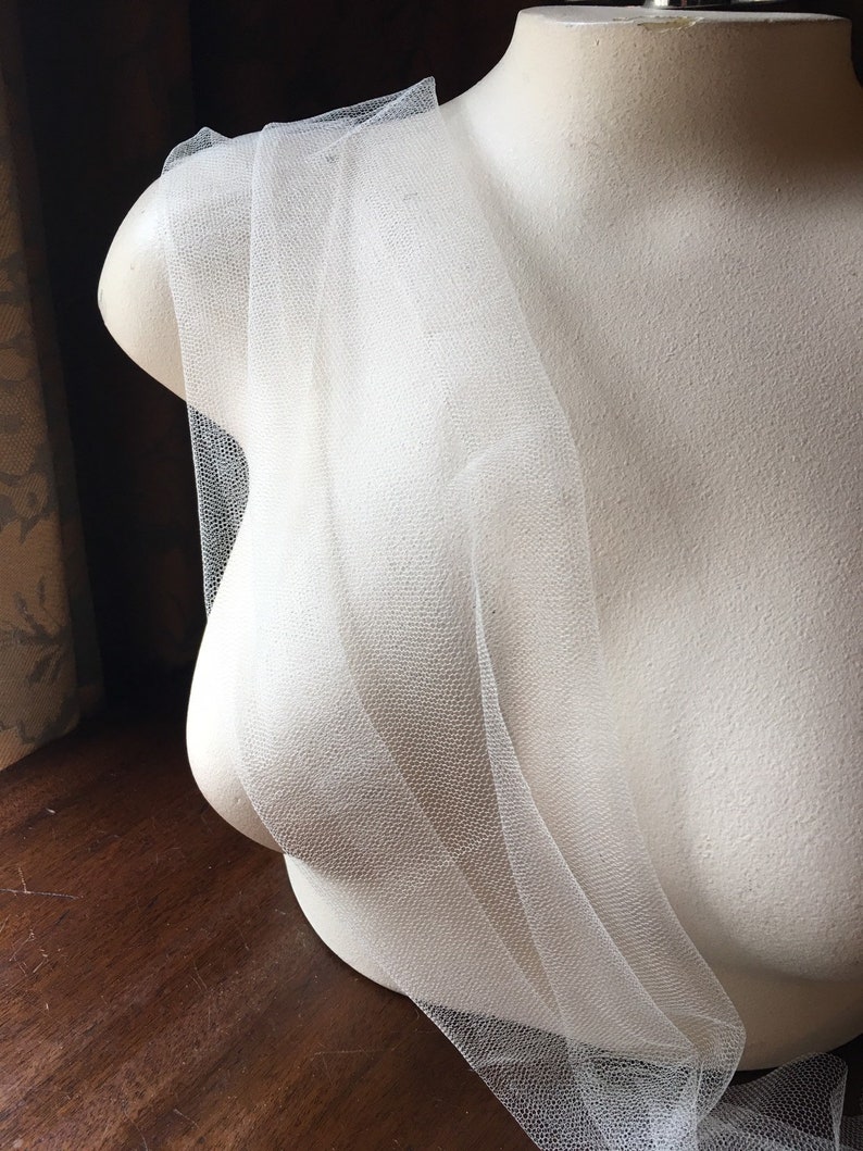 May include: A white tulle fabric draped over a mannequin bust. The tulle is sheer and delicate, with a fine mesh weave. The mannequin is a neutral off-white color, and the background is a dark wood surface.