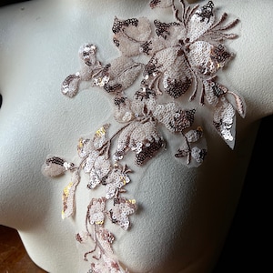 May include: A floral applique with a delicate design. The applique features a network of light pink fabric with a floral pattern. The flowers and leaves are embellished with rose gold sequins, creating a shimmering effect. The applique is displayed on a mannequin.