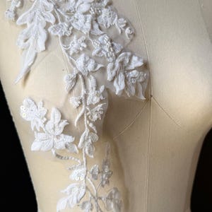 May include: A white floral lace applique featuring intricate embroidery, delicate fabric petals, and small beads on a sheer mesh backing. The design displays a trailing vine pattern with leaves and blossoms, attached to a beige dress form.