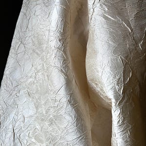 May include: Close-up of a cream-coloured fabric with a textured, wrinkled surface. The fabric appears to be draped, with folds and shadows creating depth. The material has a subtle sheen, reflecting light.