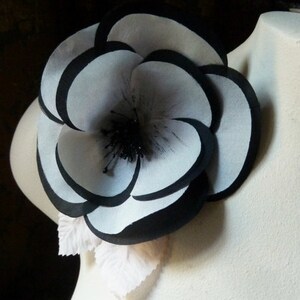 May include: A large white flower with black edges, made of fabric, with a black center. The flower is attached to a white fabric base.