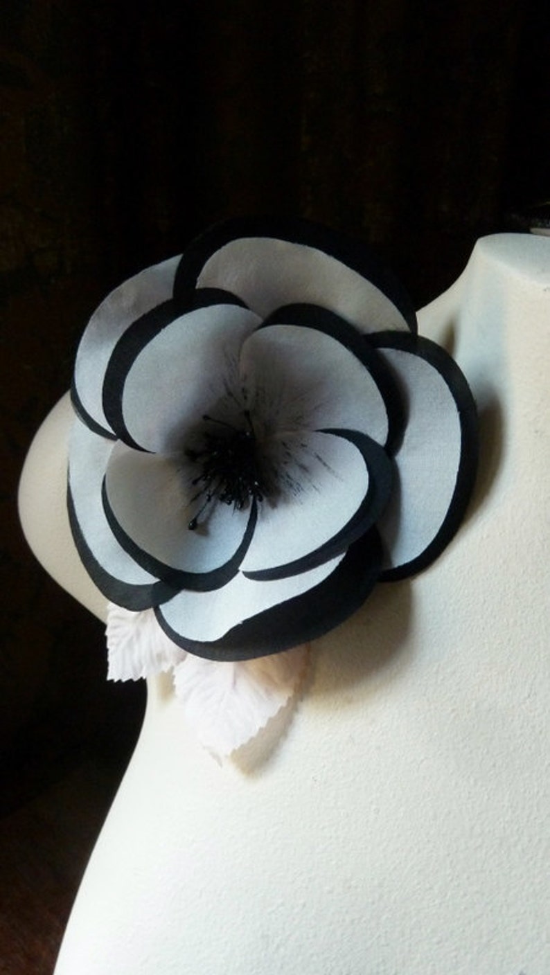 May include: A large white flower with black edges, made of fabric, with a black center. The flower is attached to a white fabric base.