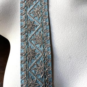 May include: Close-up of a decorative strap with intricate silver embroidery on a light blue fabric. The design features floral and geometric patterns, with a beaded border. The strap is likely part of a garment or accessory.