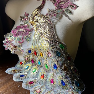May include: A gold and iridescent peacock applique with colorful rhinestones and sequins. The peacock is standing on a floral design with pink and green accents.