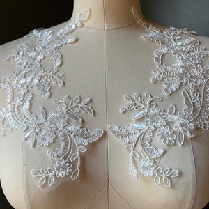 May include: Two white lace appliques with floral and leaf designs are displayed on a mannequin. The appliques feature intricate details, including small flowers and delicate, curving lines. The lace is a bright white color.