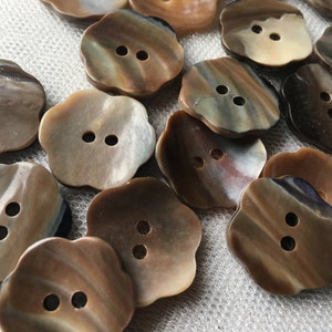 May include: A collection of flower-shaped buttons with two holes each. The buttons have a pearlescent finish with shades of brown, tan, and cream. The buttons are arranged on a textured, off-white surface.