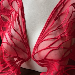 May include: A vibrant red, sheer garment with an intricate, vein-like pattern. The fabric is draped over a mannequin, showcasing the delicate, openwork design. The garment's edges are finished with a matching red trim.
