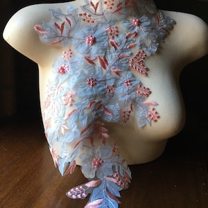 Może przedstawiać: Dekoracyjna aplikacja kwiatowa z jasnoniebieskimi, różowymi i brzoskwiniowymi akcentami. Projekt zawiera haftowane kwiaty, liście i akcenty z koralików. Aplikacja jest prezentowana na manekinie, podkreślając szczegóły.