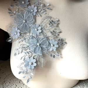 May include: A silver lace applique with delicate floral designs and pearl accents. The applique features a cluster of flowers with intricate details and a soft, shimmery finish.
