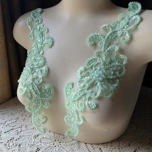 Op de afbeelding: Twee symmetrische mintgroene kanten applicaties met ingewikkeld bloemenborduurwerk, fijn kraalwerk en lovertjes. Deze decoratieve stukken zijn ontworpen met krullende patronen en worden getoond op een mannequin-torso in een neutrale kleur.