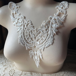 May include: A white lace applique with a delicate floral design. The applique is shaped like a V and has a scalloped edge. It is perfect for adding a touch of elegance to any garment.