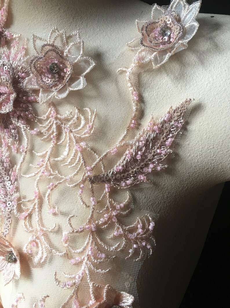 May include: A delicate lace applique with pink and gold embellishments. The applique features floral designs with beaded accents and sequins.