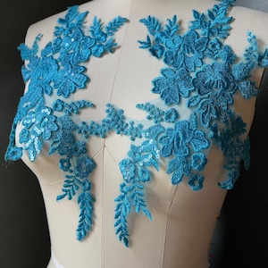 May include: Two pieces of turquoise floral lace applique. The lace features detailed flower and leaf patterns, embellished with sequins. The appliques are displayed on a mannequin, showcasing their potential use in garment design and embellishment.