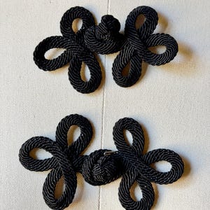 May include: Two black frog closures made of braided cord. Each fastener features a central knot flanked by two symmetrical, petal-like loops on either side, creating a decorative, floral-inspired shape for garment fastening.