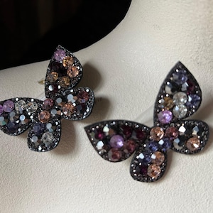 May include: A pair of butterfly-shaped earrings with a black metal base and colorful rhinestones. The earrings feature a mix of purple, pink, and brown rhinestones.