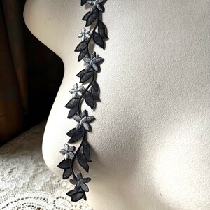 May include: A black floral applique with silver accents. The applique features a vine-like design with black leaves and silver-centered flowers. The decorative trim is displayed on a mannequin, showcasing its potential use in fashion or crafts.