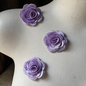 Puede incluir: Tres adornos de tela de color lavanda en forma de rosa sobre un maniquí blanco. Las rosas están superpuestas y tienen una textura ligeramente rizada.