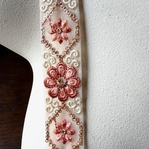 Può includere: Un nastro decorativo con una base in rete beige trasparente e motivi floreali ricamati in tonalità rosa antico e crema. Il design presenta motivi geometrici esagonali ripetuti delineati con filo metallico, alternati a petali cuciti.