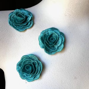 Puede incluir: Tres apliques de rosas bordadas en color teal. Los adornos florales están dispuestos sobre una superficie blanca. Cada rosa tiene múltiples capas de pétalos, creando un efecto texturizado y dimensional. El color teal tiene un ligero brillo.