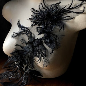 May include: Black floral lace applique with detailed embroidery. The applique features a layered design with floral and leaf patterns. The lace is attached to a sheer mesh backing.
