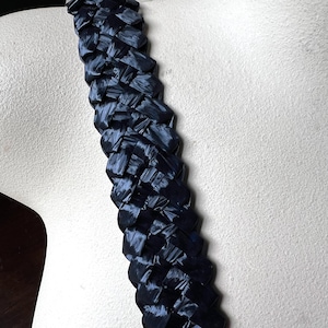 May include: A close-up of a woven, navy blue strap. The strap is made of a shiny, ribbon-like material, and is displayed on a mannequin. The weave is tight and the strap appears to be sturdy. The strap is a dark blue colour.