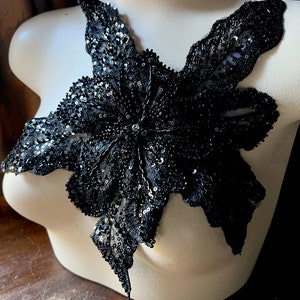 Pode incluir: Aplique floral preto com lantejoulas e contas, exibido em um manequim branco. O aplique é uma flor grande e estilizada com múltiplas pétalas, ideal para embelezar roupas ou acessórios. O design intrincado é visível no close-up.