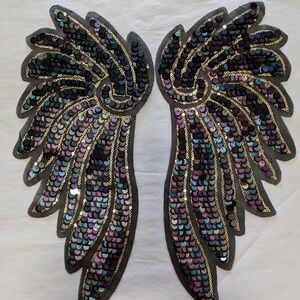 May include: A pair of iridescent black and gold sequined angel wings. The wings are made of a mesh fabric and are decorated with sequins in a variety of colors, including black, gold, purple, and green.