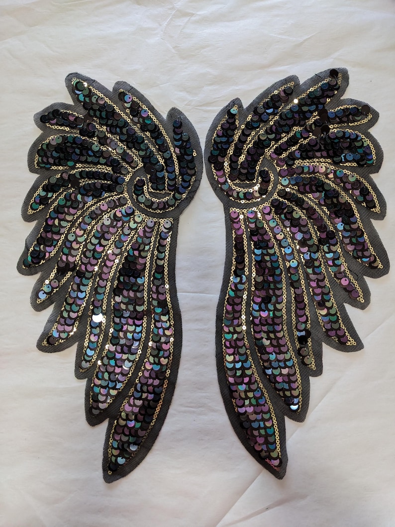 May include: A pair of iridescent black and gold sequined angel wings. The wings are made of a mesh fabric and are decorated with sequins in a variety of colors, including black, gold, purple, and green.