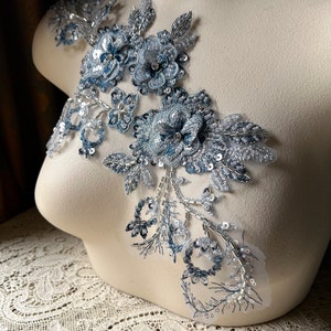 May include: A delicate, floral applique in shades of silver and blue, featuring intricate beadwork and sequins. The design includes multiple flower shapes and leaf accents, creating a three-dimensional effect. The applique is displayed on a mannequin.