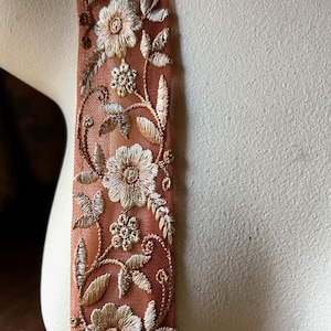 May include: Close-up of a decorative ribbon with an embroidered floral design. The ribbon is a muted orange-brown color, with detailed embroidery in gold and off-white thread. The floral pattern includes flowers, leaves, and vines, creating a delicate and ornate look.
