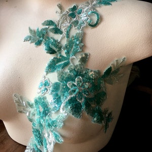 May include: A teal and silver sequined floral applique with a delicate lace design. This embellishment is perfect for adding a touch of sparkle and elegance to clothing, accessories, or home decor projects.