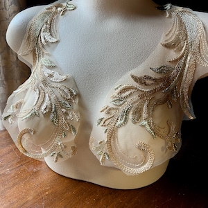 Op de afbeelding: Twee gouden en groene geborduurde kant applicaties met een bloemenmotief op een witte mannequin.