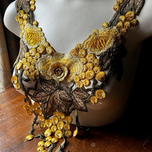 May include: An embroidered floral applique in shades of yellow, gold, and brown, featuring a rose and grape cluster design. The applique is displayed on a mannequin, showcasing the intricate details of the fabric and stitching.