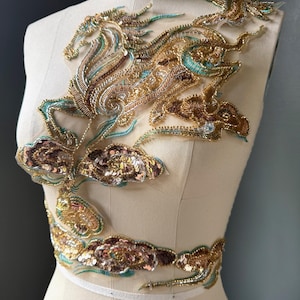 May include: An ornate, gold-toned beaded applique featuring a mythical horse design with flowing mane and tail. The design incorporates teal and bronze accents, with sequined cloud-like embellishments. The applique is displayed on a mannequin.