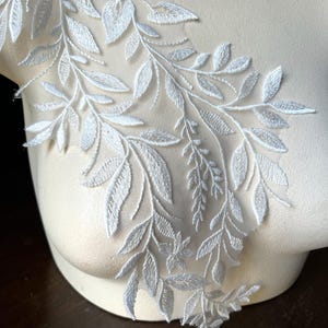 Puede incluir: Aplicación de encaje floral blanco con un delicado diseño de hojas y vides. La aplicación está hecha de una tela suave y transparente, ideal para adornar prendas o accesorios. Los intrincados detalles y el diseño elegante lo convierten en una hermosa adición a cualquier proyecto.