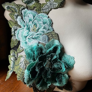 May include: A pair of teal and green embroidered floral appliques. The appliques feature two large flowers with detailed stitching and smaller green leaves.