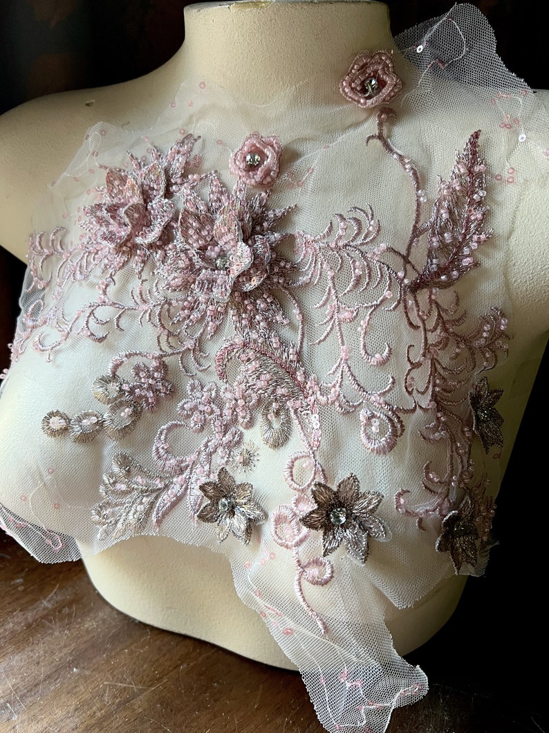 May include: Pink and brown floral lace applique with beaded details. The applique is on a sheer white mesh fabric.