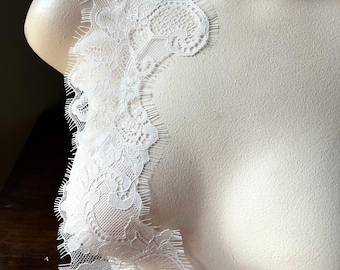 May include: A delicate white lace trim is draped over a mannequin. The lace features intricate floral and geometric patterns with a scalloped edge. The trim is likely used for embellishing garments or crafts.