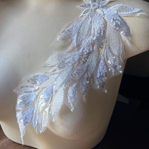 May include: White lace applique with silver sequins in a floral design. The applique features a large flower with smaller leaves and petals.