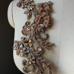 May include: A decorative appliqué featuring intricate embroidery, light brown beads, and clear faceted crystals arranged in a floral and leaf pattern on a sheer mesh base. The design includes layered fabric petals and metallic thread accents.
