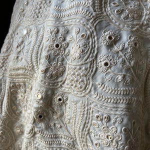 May include: Close-up of cream-coloured fabric with detailed embroidery and small mirror embellishments. The design includes floral motifs, curved lines, and fine stitching, giving a textured and decorative look. The fabric seems to be a delicate textile.