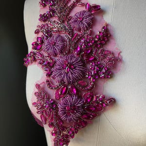 May include: A magenta floral applique with beaded and rhinestone embellishments. The design features three large, beaded flower clusters and smaller floral and leaf accents. The applique is set on a sheer, pink mesh backing.