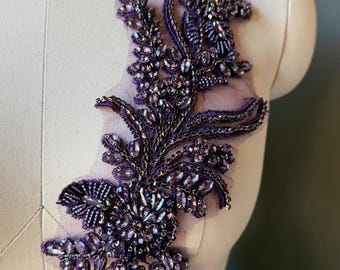 May include: A dark purple floral applique featuring intricate beadwork and iridescent crystals on a sheer mesh backing. The design showcases a central flower motif surrounded by leafy vine patterns, all embellished with shimmering purple stones.
