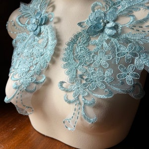 May include: Two light blue lace appliques with floral designs and pearl embellishments. The appliques are on a white mannequin bust.